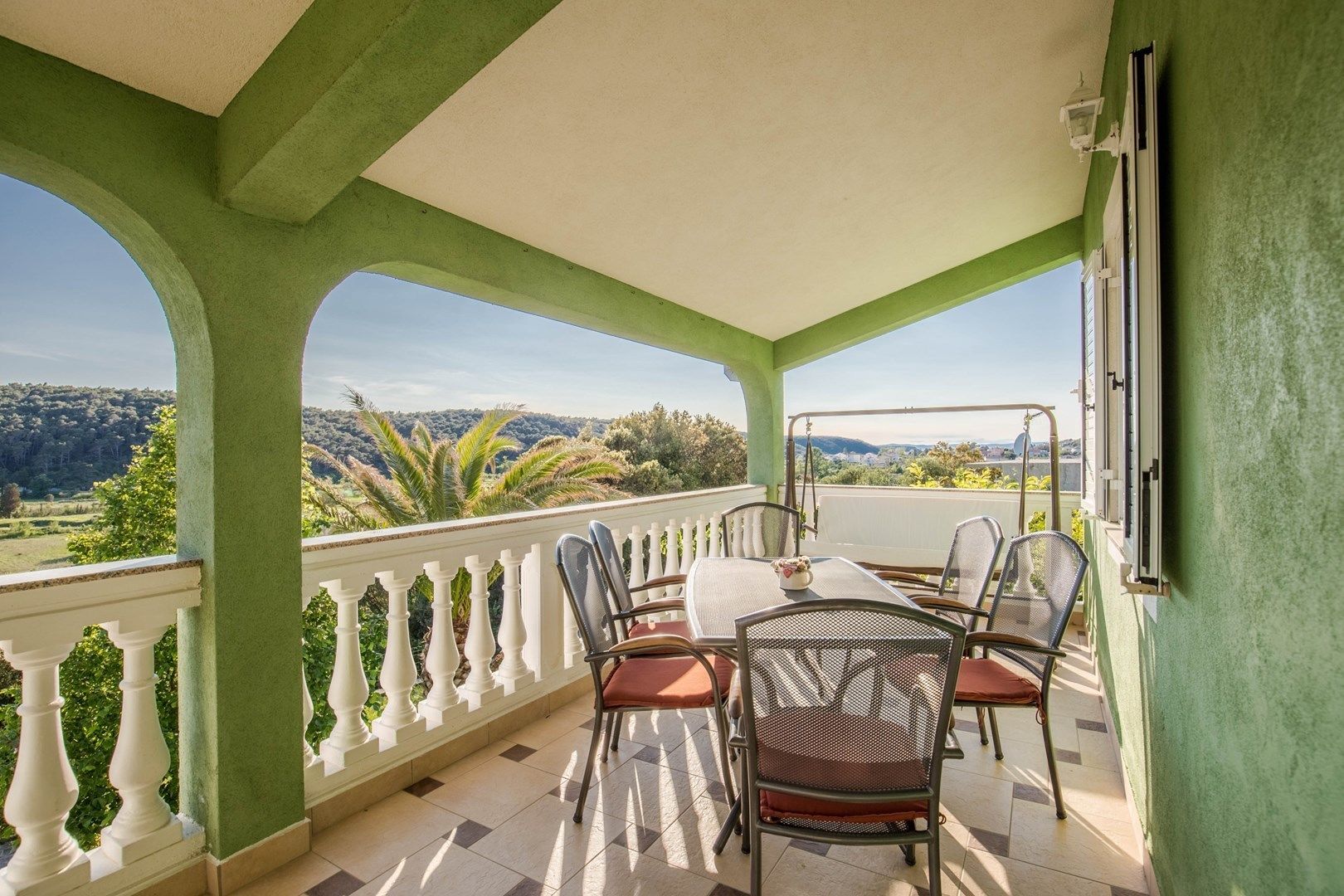 A balcony with a table and chairs, white balustrade, and a view of a hilly landscape.