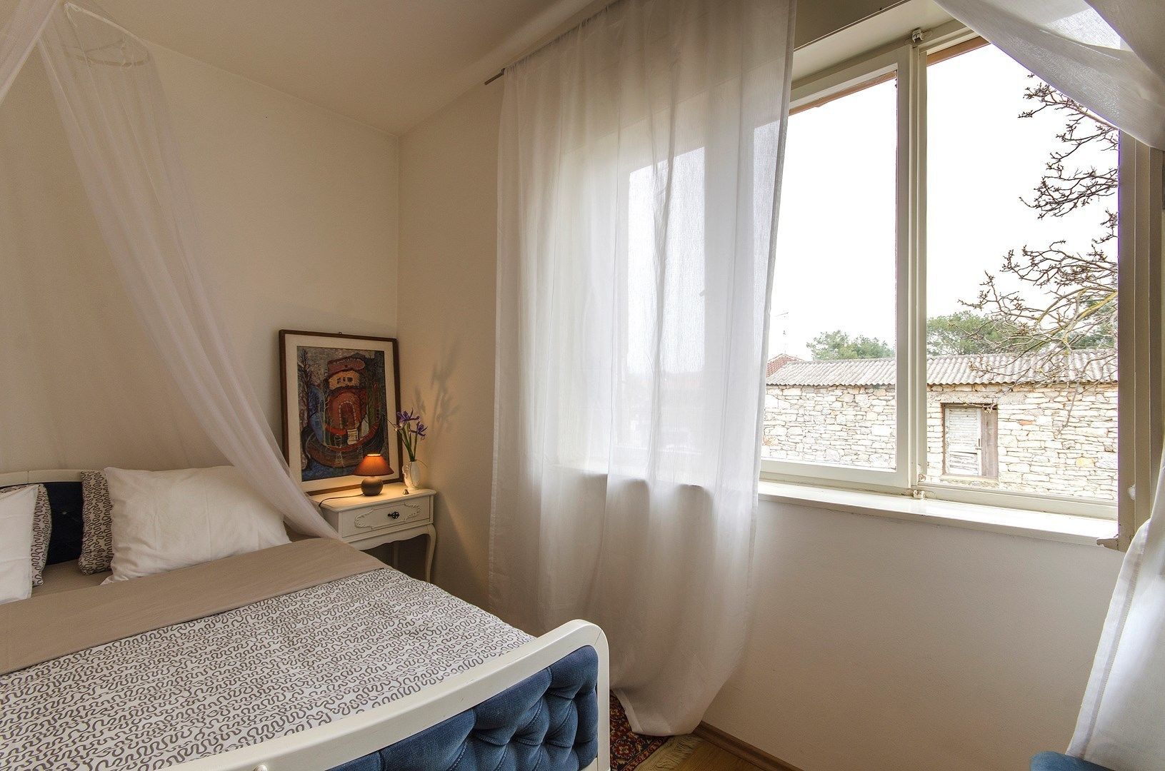 A bedroom with a bed, a bedside table with a lamp, a framed picture, and a window with sheer curtains.