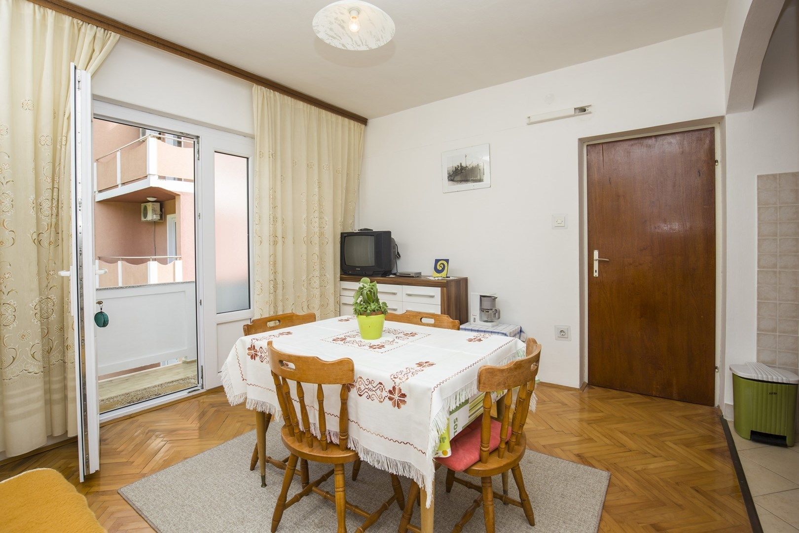 Un comedor con una mesa preparada para cuatro personas, sillas de madera, una planta en maceta y un televisor sobre una mesita.