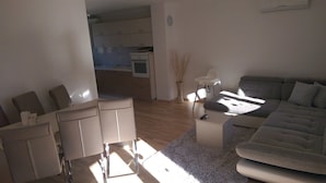Un salon moderne avec un canapé, des fauteuils et une petite table. On aperçoit une cuisine au fond.