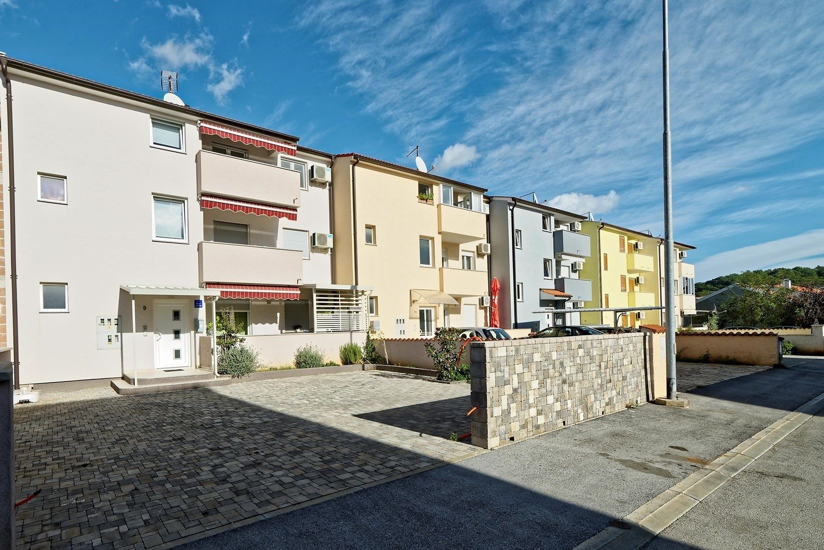 A row of multi-story residential buildings with balconies, a paved courtyard, and a stone wall.