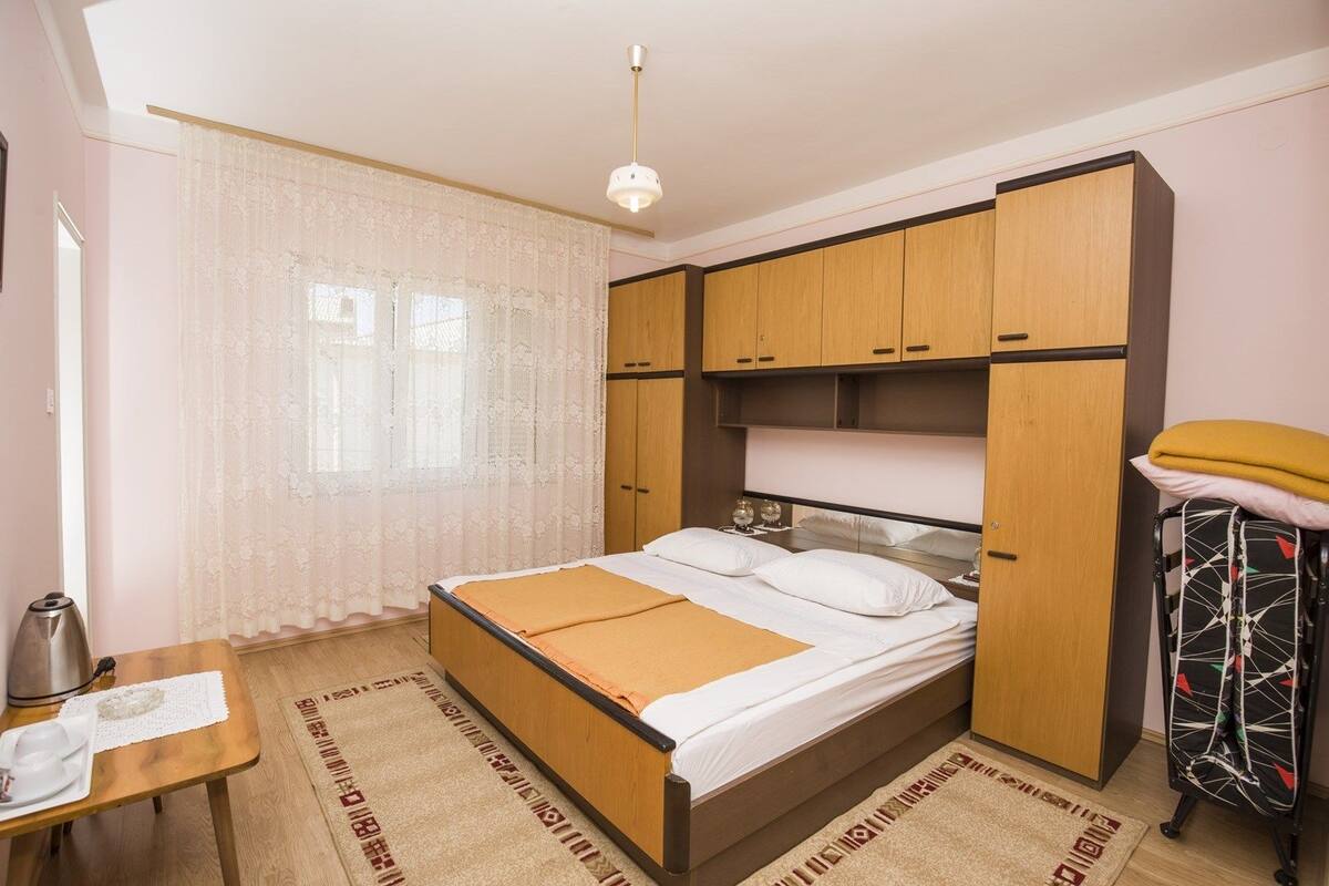 Quarto com cama, guarda-roupa de madeira, mesa com chaleira e janela com cortinas leves.
