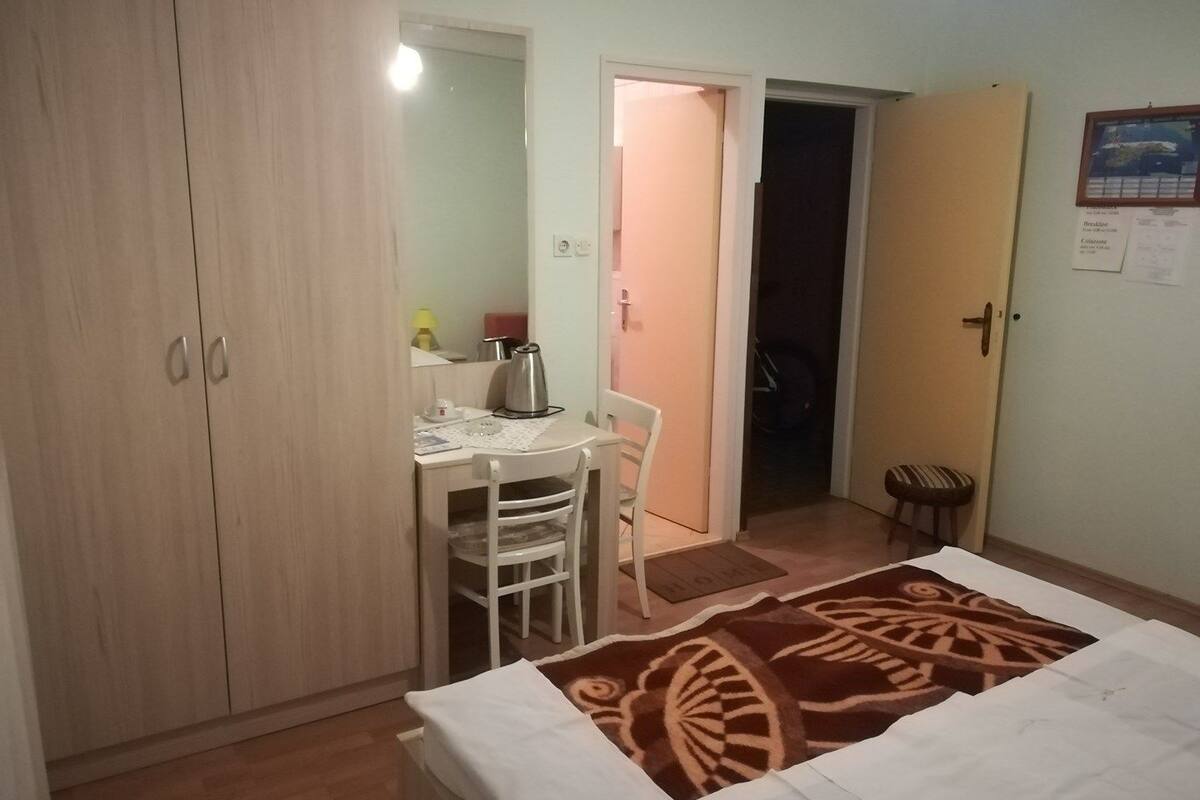 Um quarto pequeno e bem iluminado, com uma cama de solteiro, uma escrivaninha com cadeira, um guarda-roupa e uma porta que dá para outro cômodo.