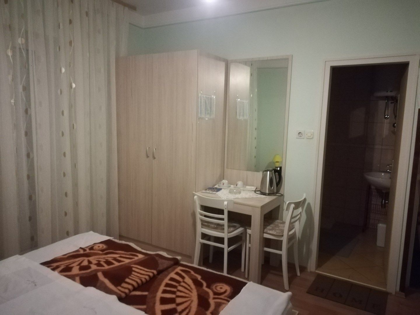 A bedroom with a bed, a desk, chairs, a wardrobe, and a bathroom visible through an open door.