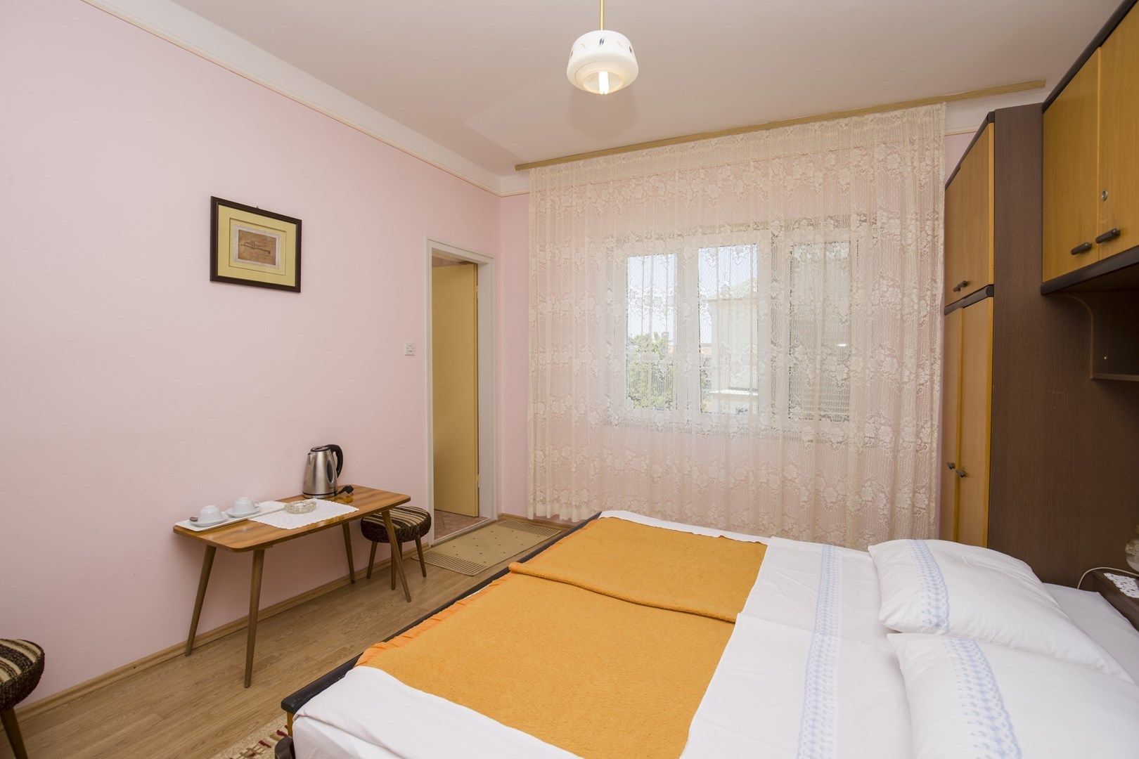 A room with a bed, a desk with a chair, a window with lace curtains, and a door.