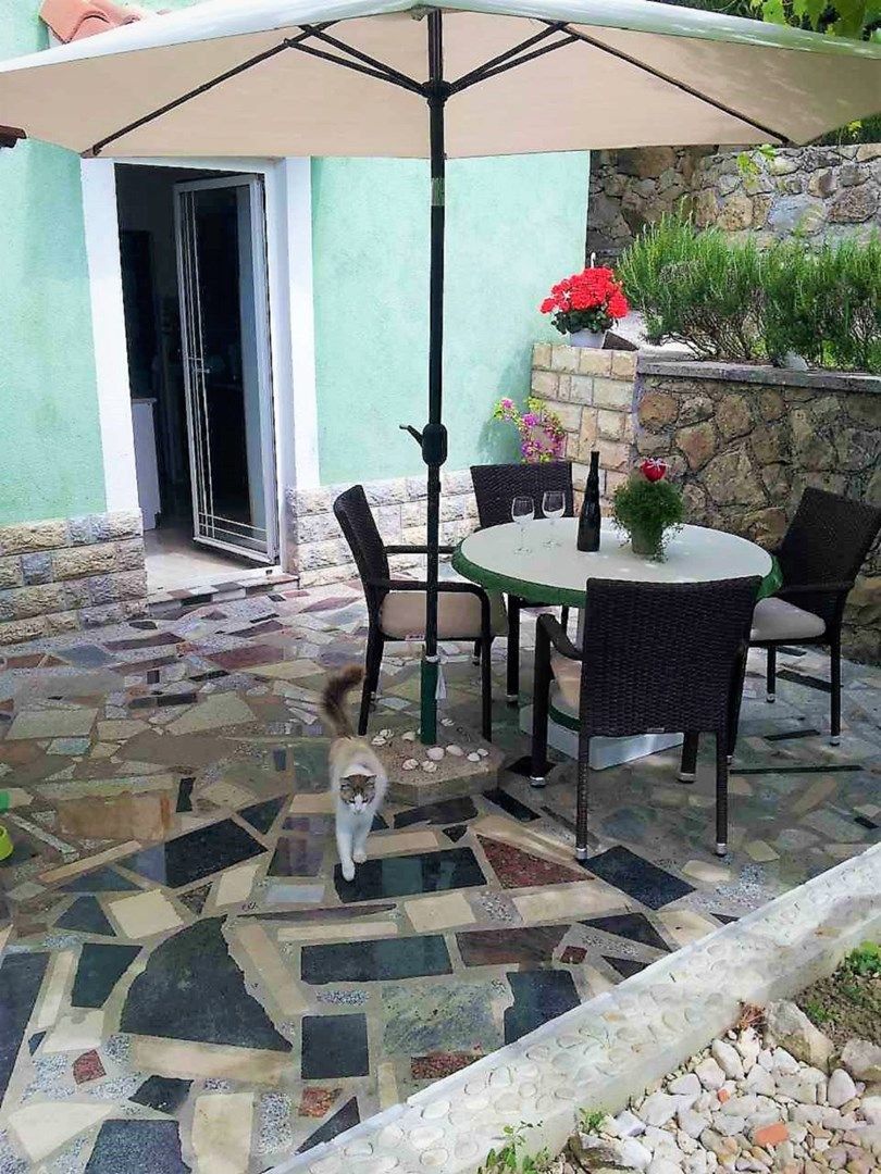 Een terras buiten met een ronde tafel en stoelen, een vloer van stenen tegels en een stenen muur met planten.