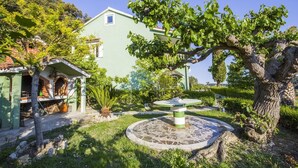 Un edificio tradicional con fachada verde, una fuente de piedra y un jardín bien cuidado con diversas plantas y un árbol.