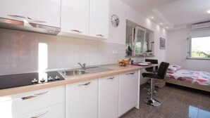 Eine moderne Küche mit weißen Schränken, einer schwarzen Arbeitsplatte und einem eingebauten Herd. Es gibt eine kleine Essecke mit einem Stuhl und ein Bett im Hintergrund.