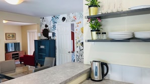 A kitchen with a countertop, a kettle, a sink, and a shelf with dishes and a potted plant.
