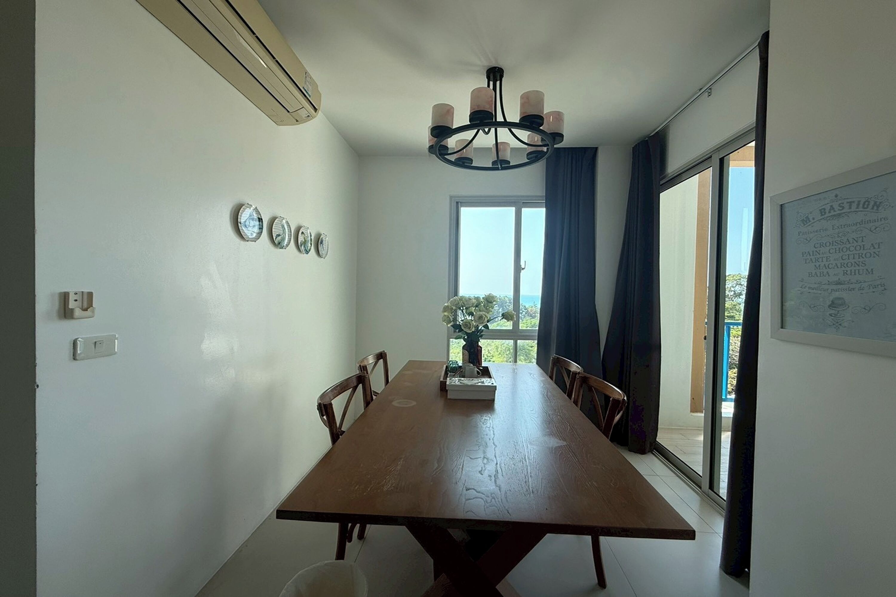 A dining area with a wooden table, chairs, a chandelier, and a view through a large window.