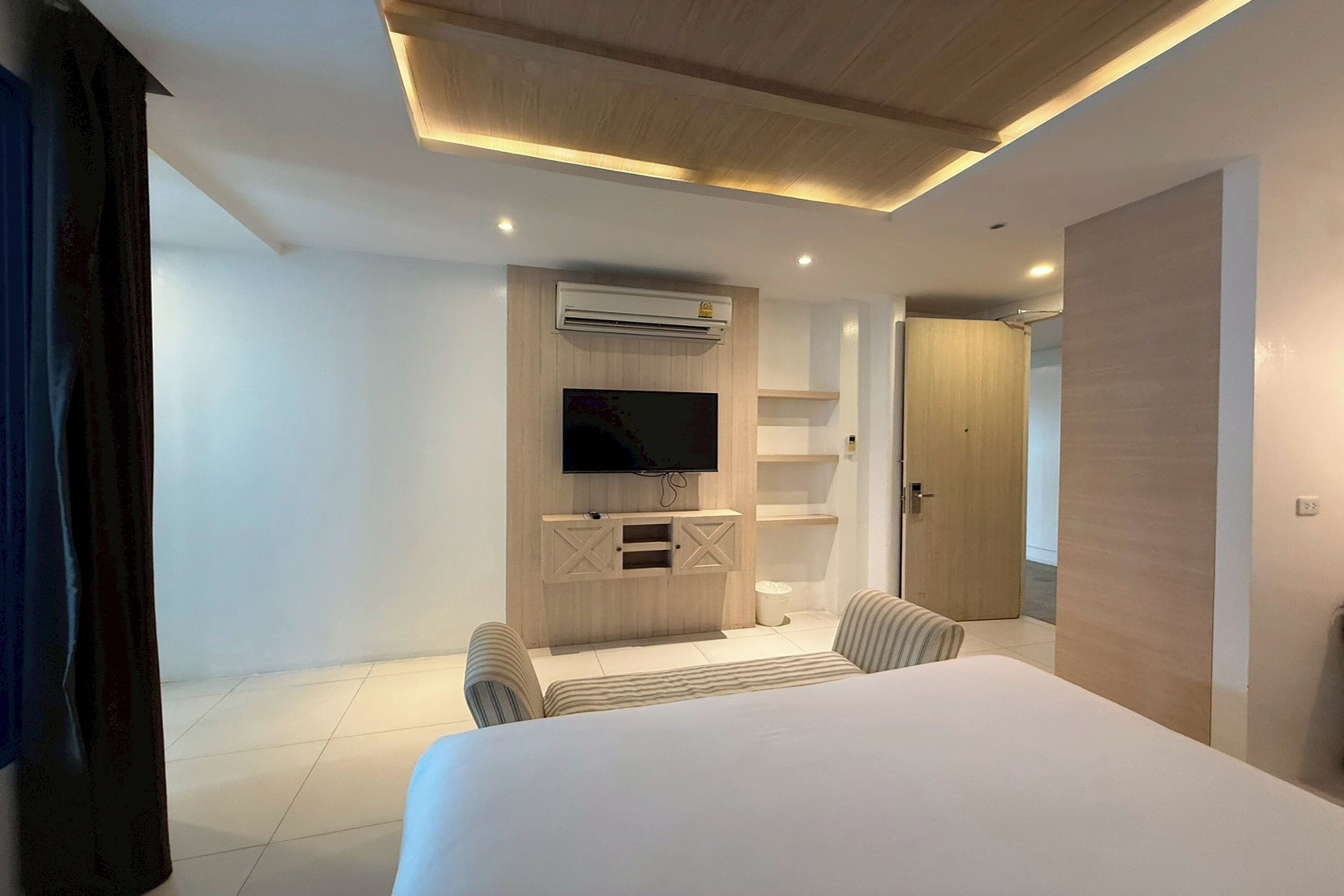 Ein modernes Hotelzimmer mit einem großen Bett, einem an einer Holzvertäfelung befestigten Fernseher, einer kleinen Sitzecke und einer Tür zu einem weiteren Raum.