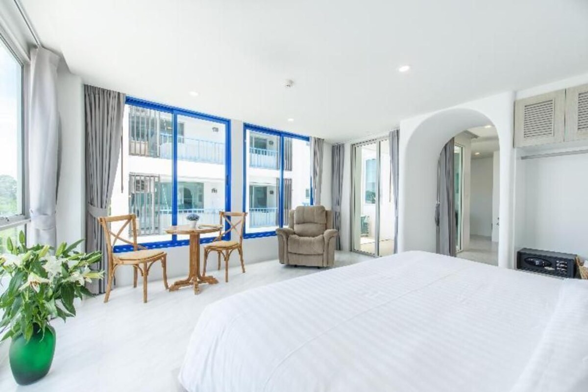Quarto de hotel moderno com uma cama grande, área de estar com cadeiras e uma mesa, um vaso de planta e janelas amplas com vista para o edifício.