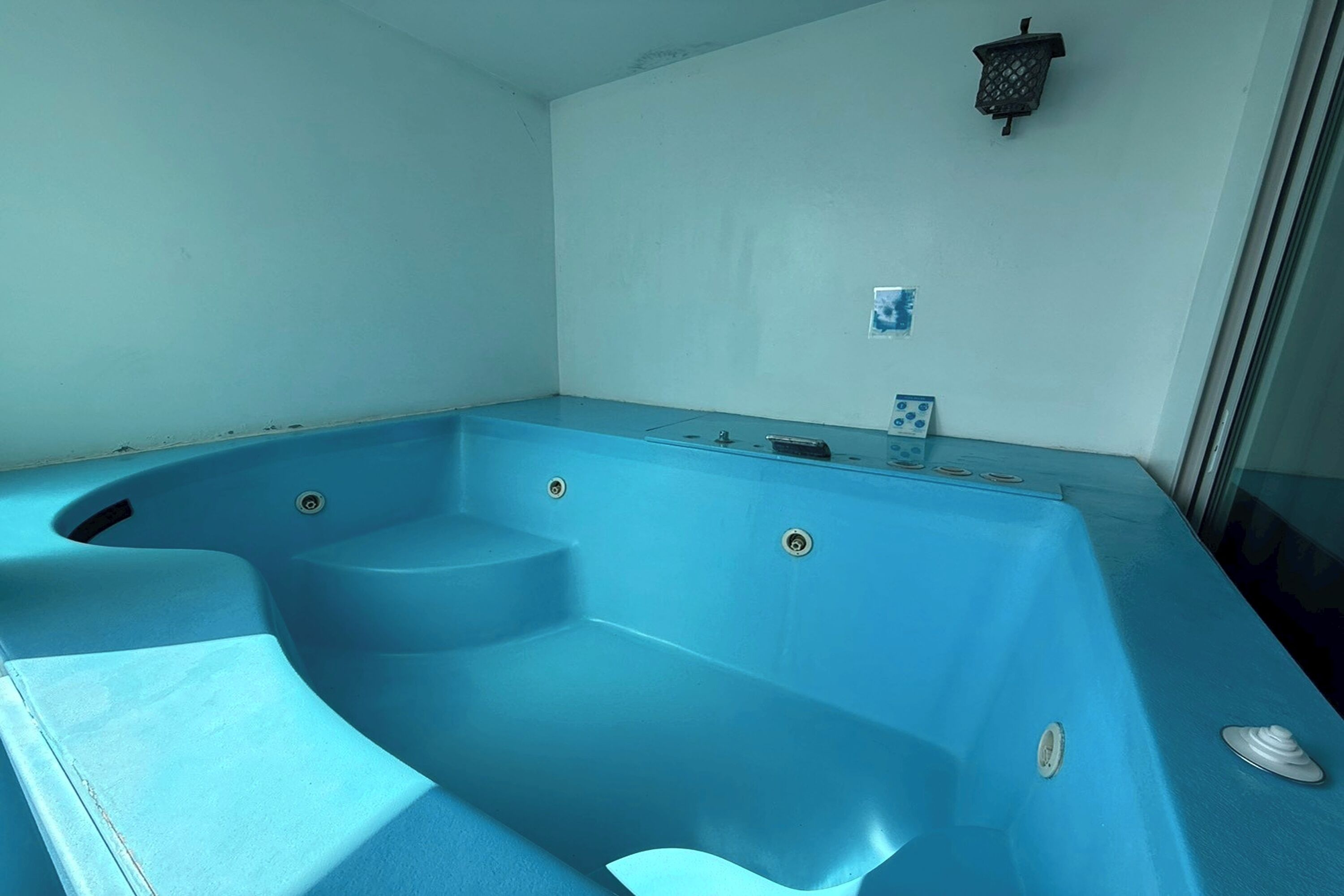 A blue jacuzzi with steps, a small wall-mounted light fixture, and a window with a view.