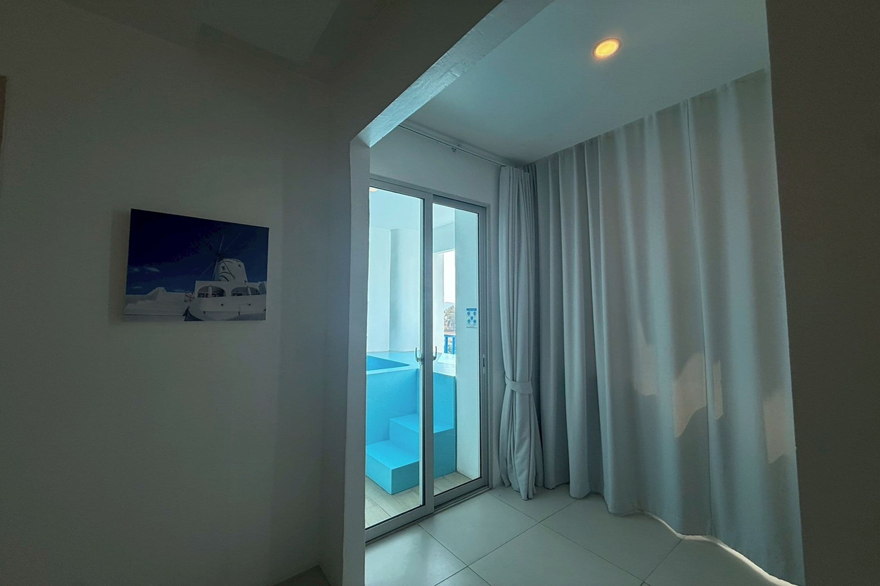A room with a large sliding glass door leading to a balcony, a white curtain on the right, and a painting on the wall.