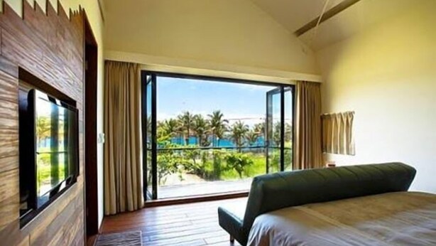 A hotel room with a large bed, a TV, and a balcony with a view of a pool and palm trees. - Coconut Beach Homestay (Beinan)