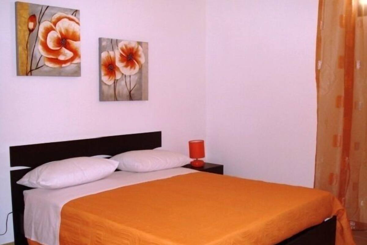 A bedroom with a bed, pillows, a nightstand, a red lamp, and two floral paintings on the wall.