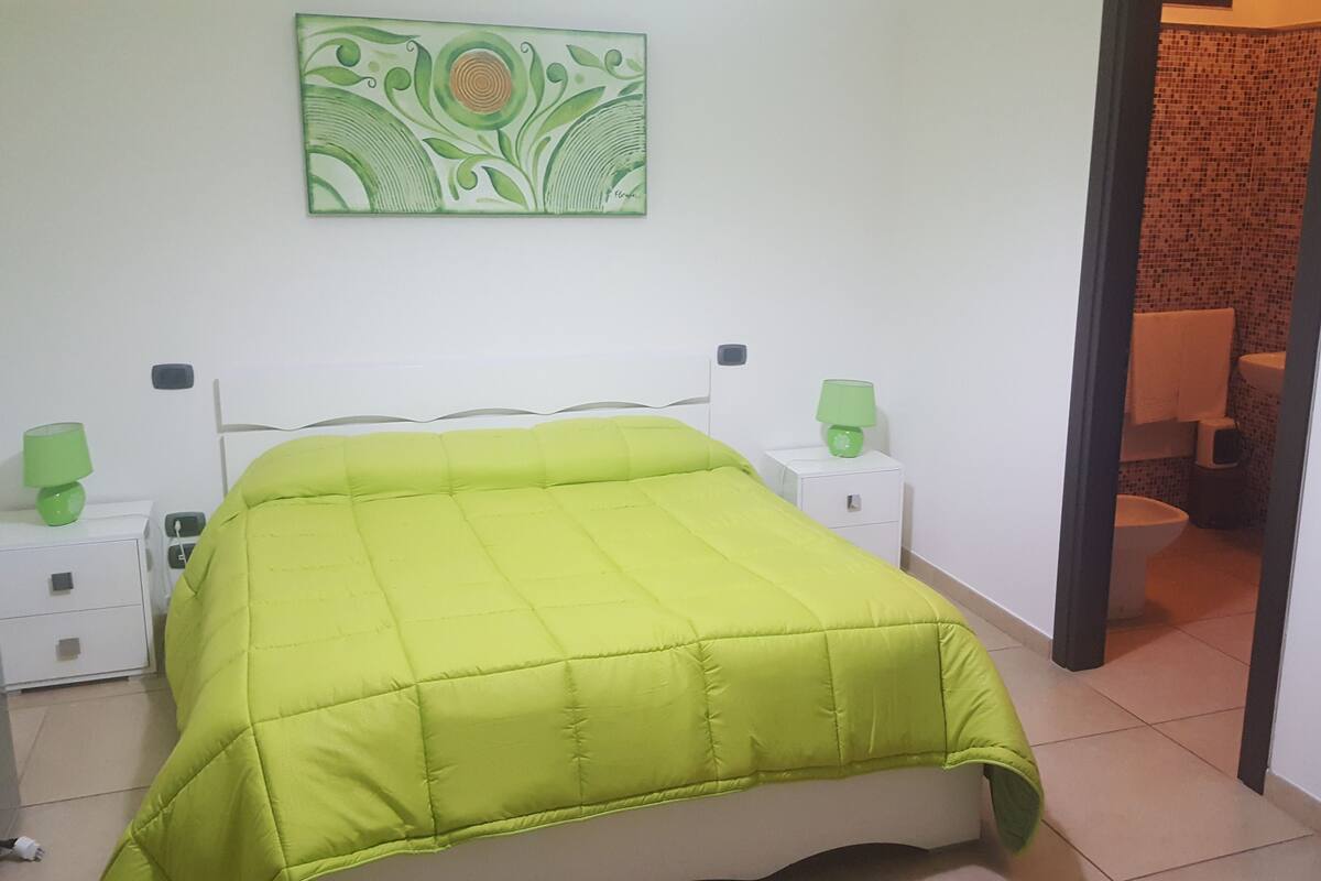 A room with a bed, two bedside tables with lamps, a wall-mounted artwork, and a bathroom visible through an open door.