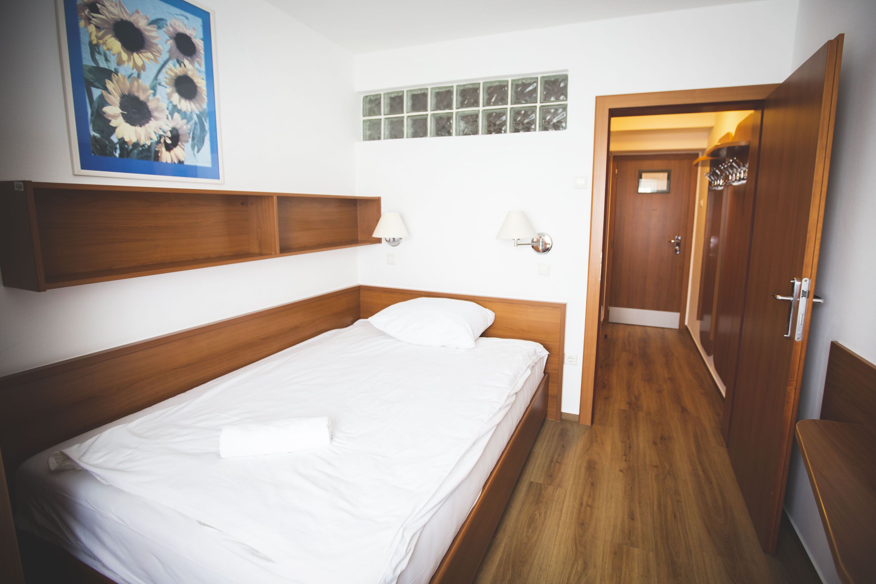 Un letto singolo con testiera in legno, un comodino, una lampada a parete e un quadro con girasoli.