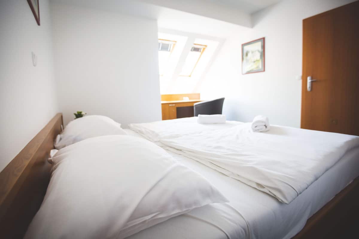 A neatly made bed with white linens in a room with a wooden headboard, a small plant, and a view of a kitchen area through an open door.