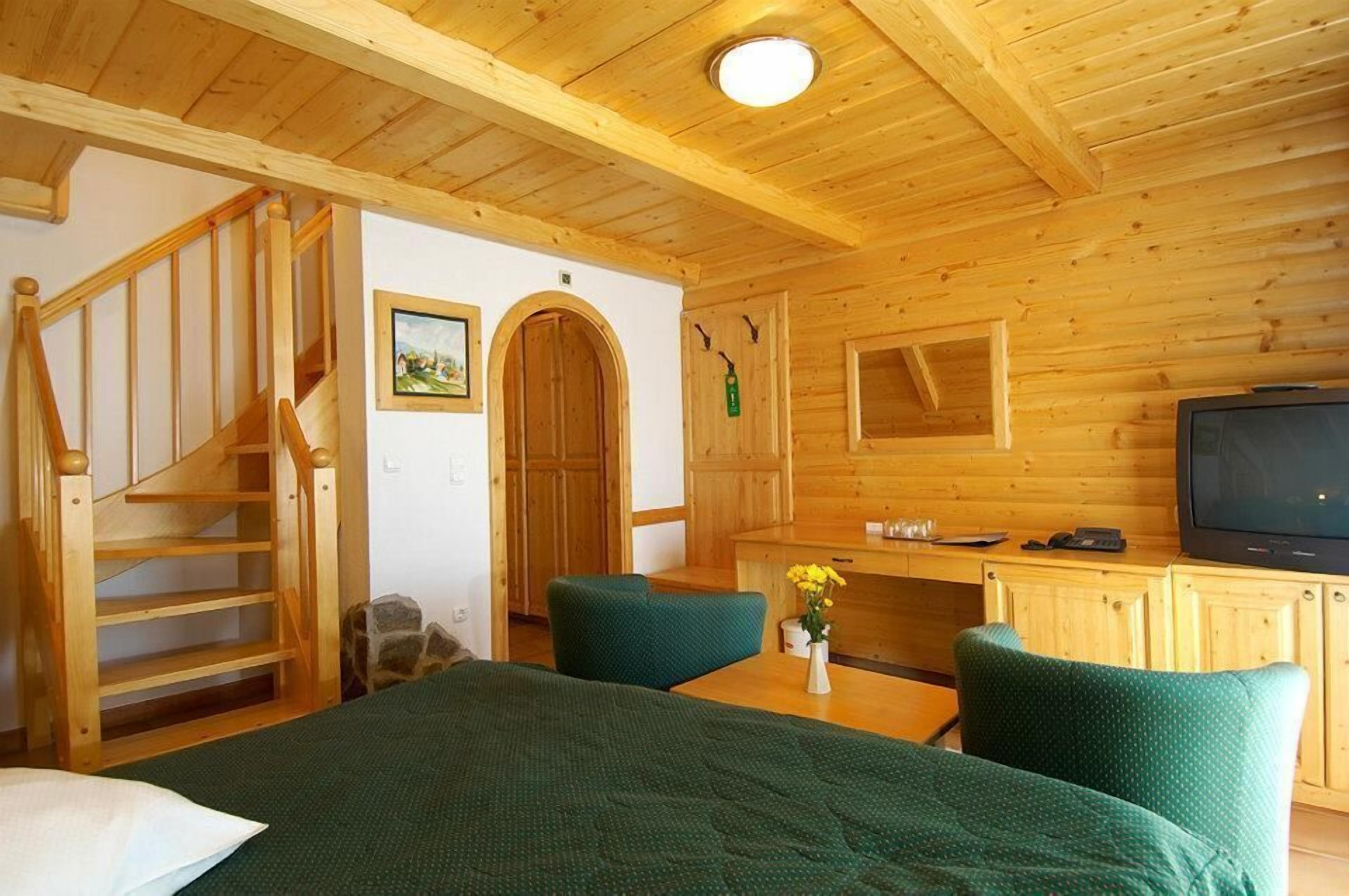 Interior de una cabaña de madera con una escalera, una cama, un escritorio con televisor y una silla verde.