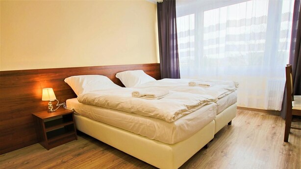 Una cama doble con cabecera de madera, una mesita de noche con lámpara y vistas al exterior a través de cortinas translúcidas.