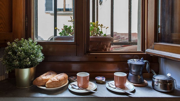 A kitchen counter with a window, a potted plant, a loaf of bread, a cup, a plate, a spoon, a coffee pot, and a small pot. - Chiasso degli Armati Apartment (Florence)