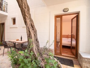 Een boom in een binnentuin met een tafel en stoelen, een deur die leidt naar een kamer met een bed, en een klein balkon met een hekwerk.