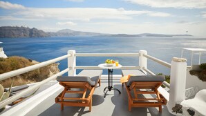 A balcony with two wooden chairs and a small table with a bowl of fruit and a glass of orange juice. The view includes a body of water and distant landforms. - Diamond Luxury Suites Collection (Santorini)
