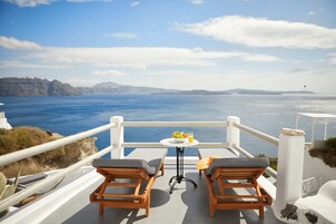A balcony with two wooden chairs and a small table with a bowl of fruit and a glass of orange juice. The view includes a body of water and distant landforms.