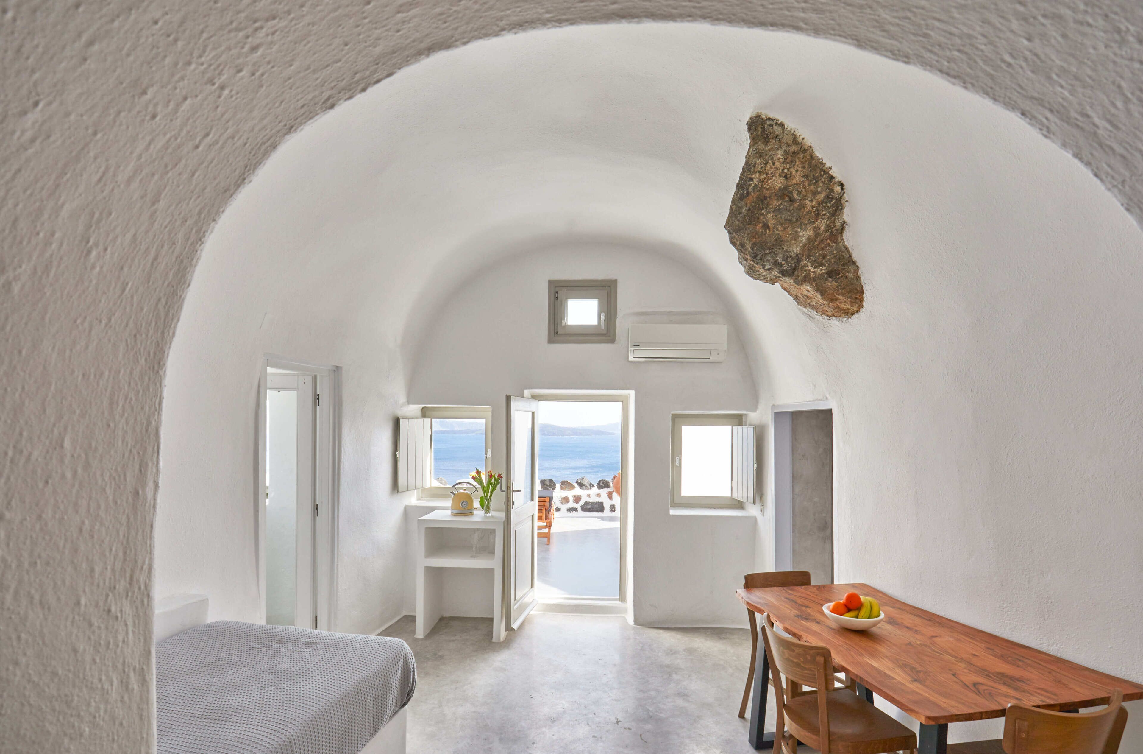 Een kamer met een wit gewelfd plafond, een houten eettafel met stoelen, een klein raam en uitzicht op zee.