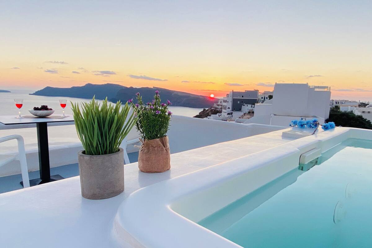 Ein Pool auf dem Dach mit Blick auf den Sonnenuntergang, zwei Kübelpflanzen und ein Tisch mit Obstschale und Gläsern.
