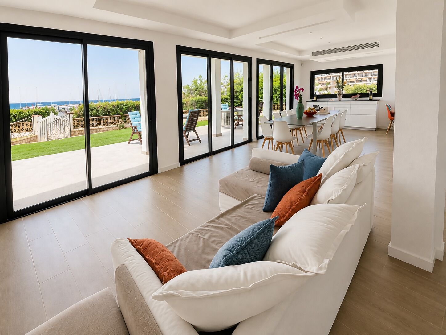 A modern living room with a large sofa, a dining area, and a view of the sea.