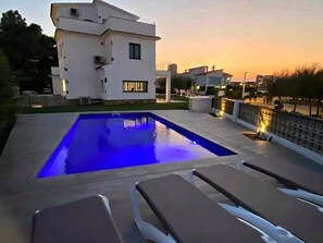 Una piscina con sillones de descanso, un edificio blanco y un atardecer de fondo.