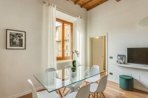 A modern living room with a glass dining table, white chairs, a green ottoman, a TV on the wall, and a window with curtains. - Apartments Florence - Luna Chic (Firenze)