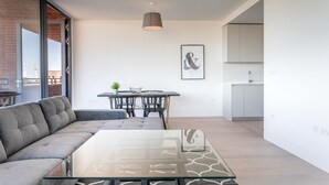 A modern living room with a glass coffee table, grey sofa, dining table with chairs, and a kitchen area with cabinets.