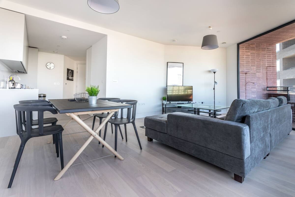 A modern living room with a grey sofa, a dining table with chairs, a TV, and a kitchen area in the background.