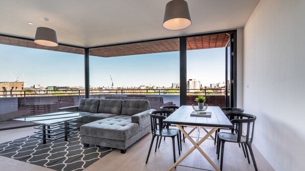 A modern living room with a sectional sofa, dining table, and chairs, large windows with a city view, and pendant lighting.