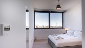 A modern hotel room with a large window, a bed, a bedside table with a lamp, and a thermostat on the wall.