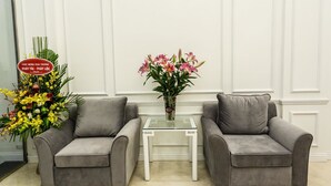 Two grey armchairs facing each other with a small white table between them, a vase of pink flowers on the table, and a large floral arrangement to the left. - Hanoi A83 Hotel (Hanoi)