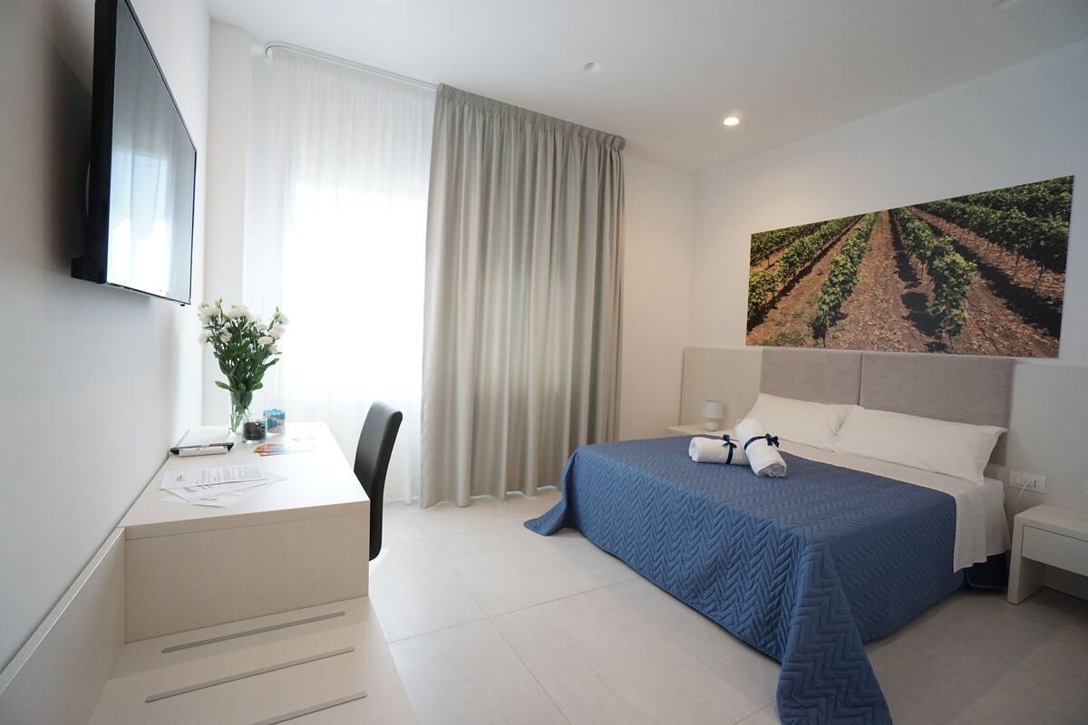 Ein Hotelzimmer mit Bett, Schreibtisch, Sessel, Fernseher und einem großen Fenster mit Vorhängen.
