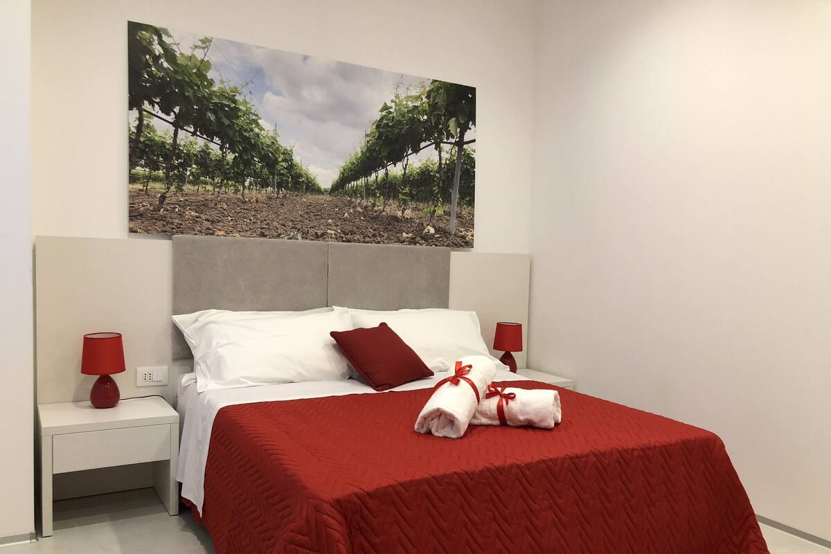 Ein Hotelzimmer mit einem großen Bett, roter Bettwäsche, zwei Nachttischen mit roten Lampen und einem Gemälde eines Weinbergs über dem Bett.