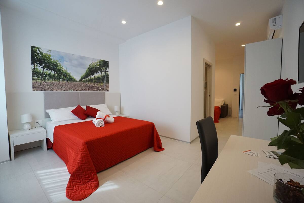 Ein Hotelzimmer mit Bett, Schreibtisch, Stuhl, Nachttisch, Lampe, einer Vase mit Blumen und einem Bild an der Wand.