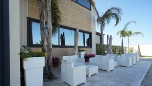 Un espace extérieur aménagé avec des meubles en osier blanc, des plantes en pot et un bâtiment doté de grandes fenêtres.