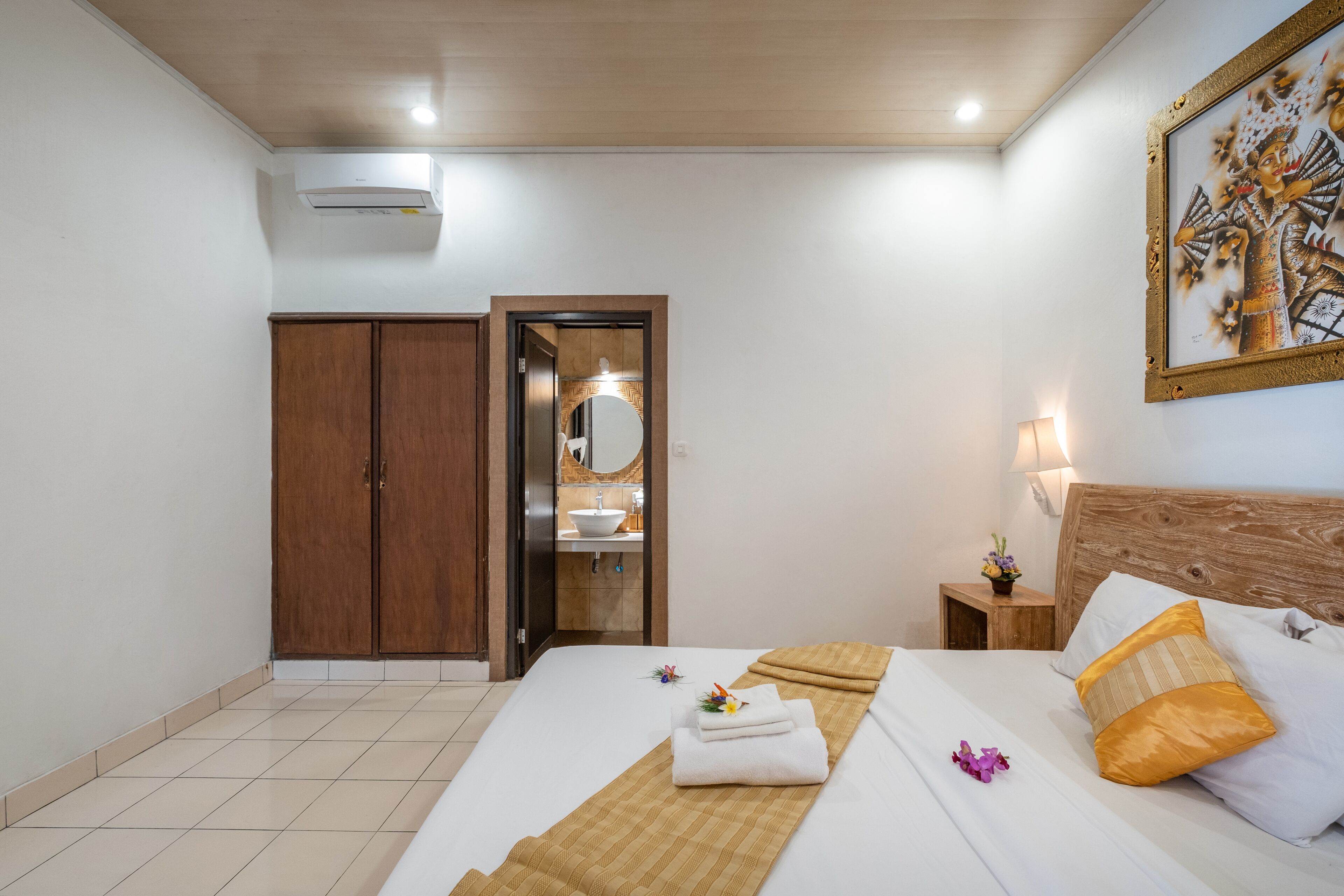 A hotel room with a bed, a wooden wardrobe, a bathroom with a sink and mirror, and a painting on the wall.