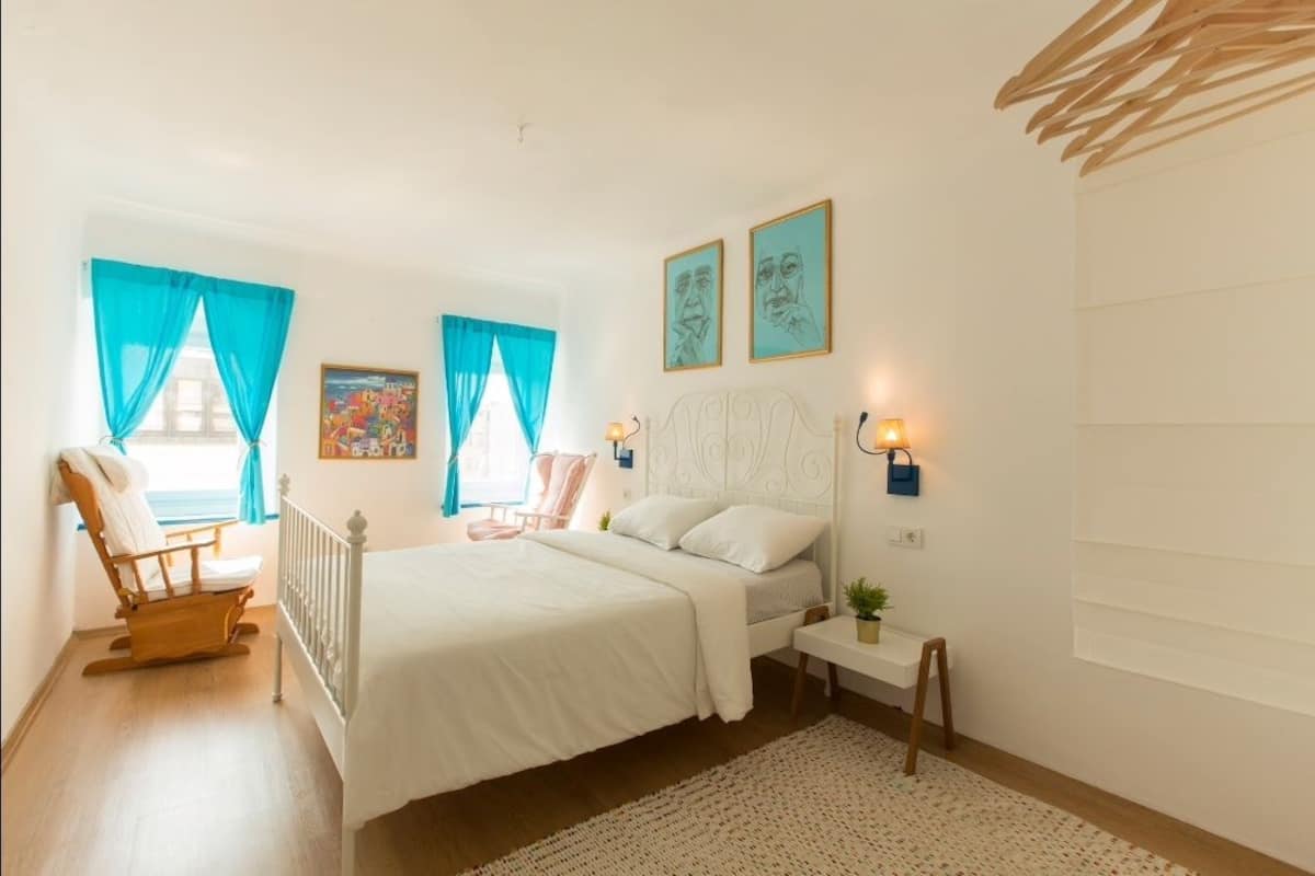 A bedroom with a bed, a chair, a bedside table, and a window with curtains.