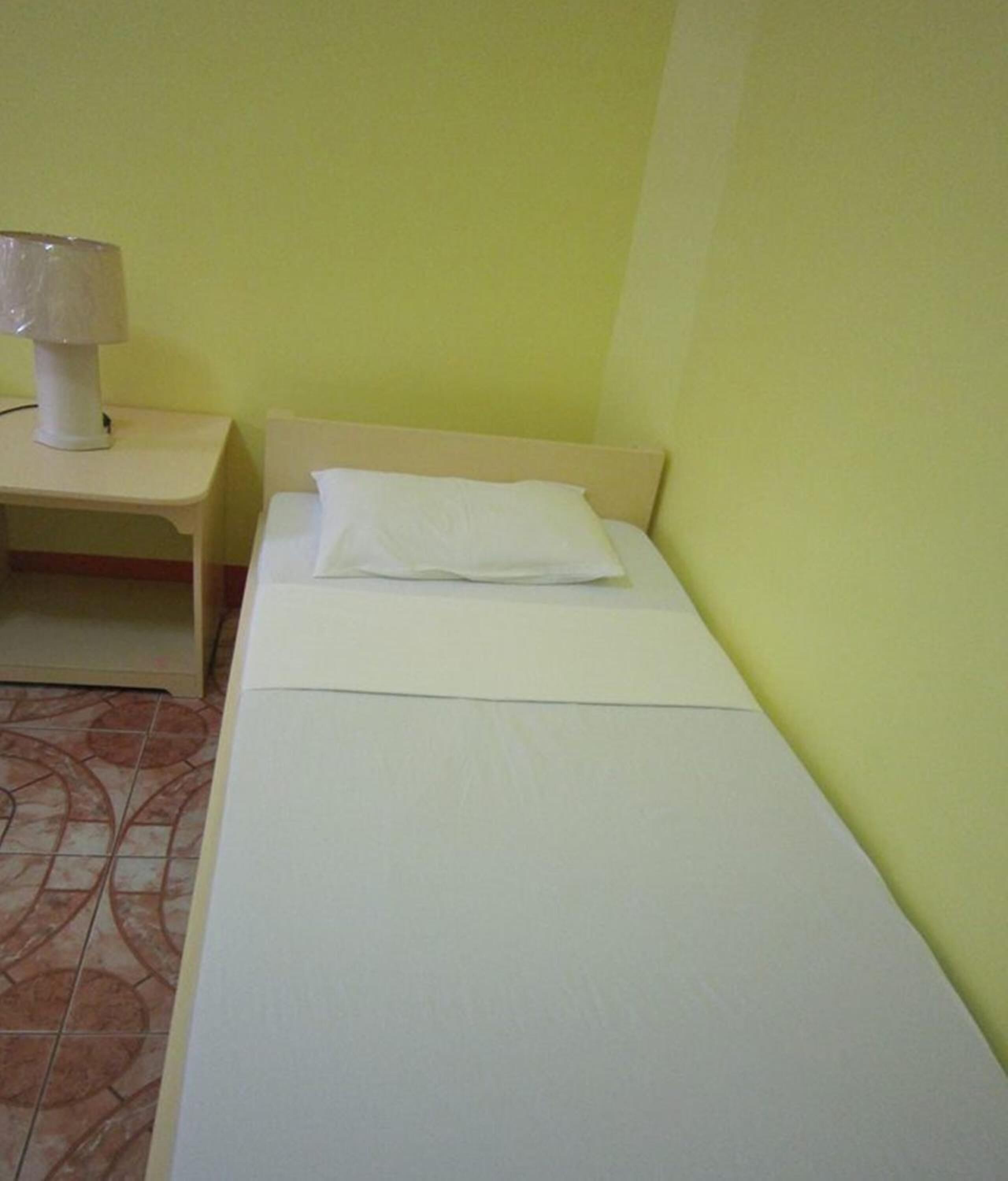 A single bed with a white mattress and pillow in a room with yellow walls and a tiled floor.