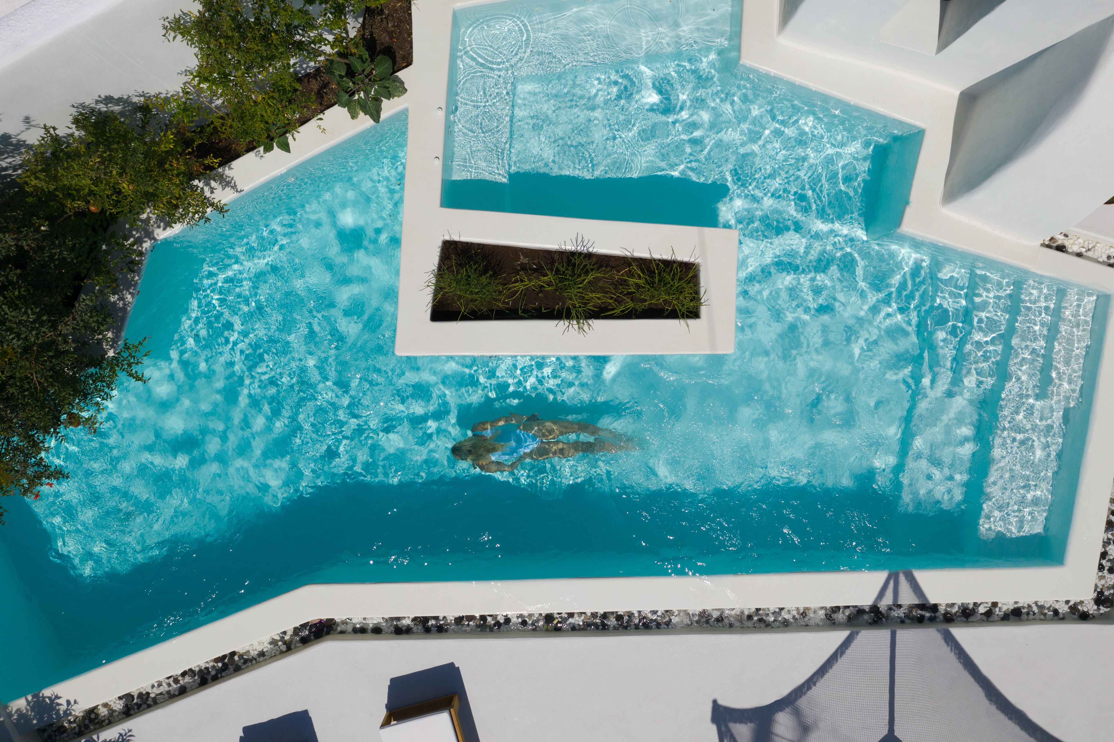 Une vue aérienne d’une piscine à l’eau d’un bleu limpide, entourée de murs blancs et d’une petite zone de végétation.
