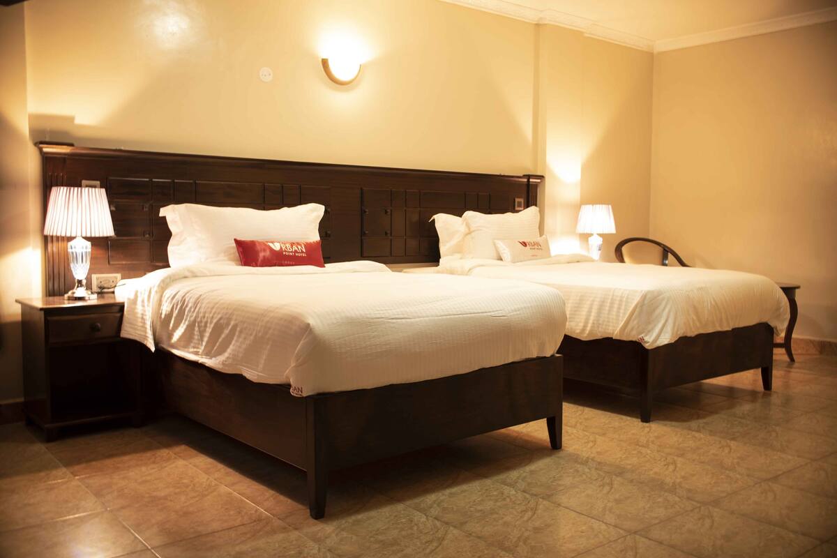 A hotel room with two beds, a nightstand with a lamp, and a wall-mounted light fixture.