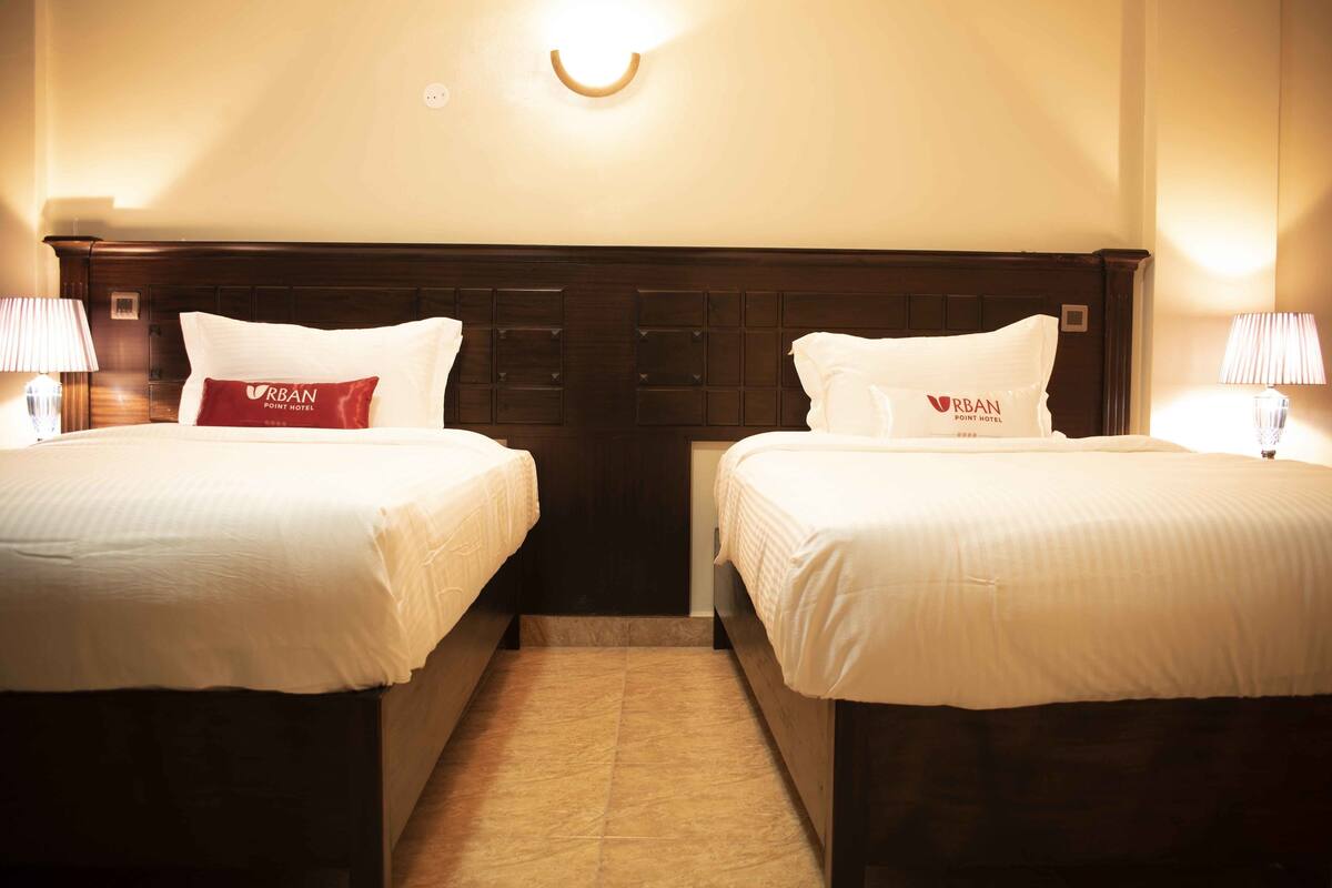A hotel room with two beds, each with a white bedspread and pillows, including a red Urban Point Hotel pillow. There are bedside lamps and a wall-mounted light fixture.