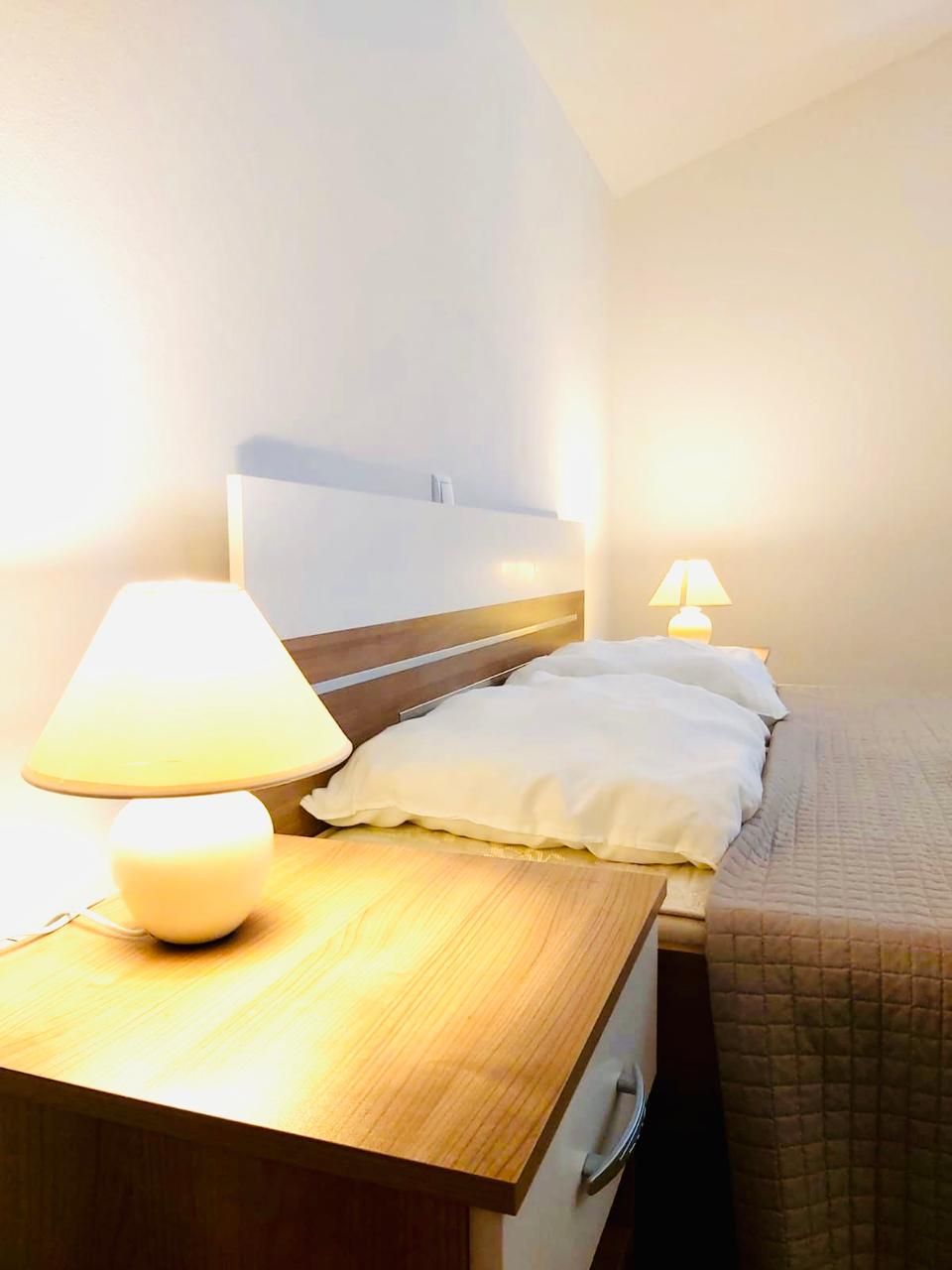 A hotel room with a wooden bedside table, a lamp, a bed with white bedding, and a headboard.